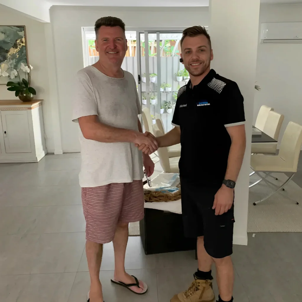 Electrician in uniform shaking hands with a happy homeowner inside a bright, modern living room, showing a successful service visit and customer satisfaction.