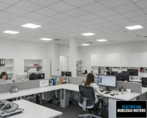 Open-plan office space with desks, computers, and employees working under grid lighting.